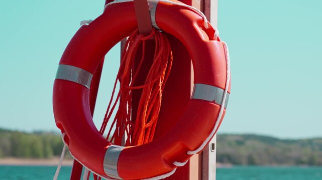 Bright red lifebuoy hangs on marina post with 112 emergency sign, rope, and metal fittings, set against calm blue water, distant shoreline, and clear summer sky