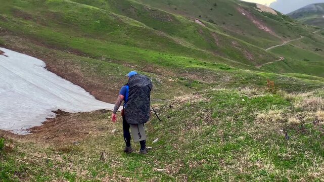 Lone hiker carrying a large trekking pack walks across remote alpine terrain with rolling green mountains and lingering snowfields in the Georgian Caucasus wilderness.