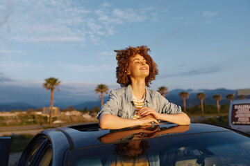 Fototapeta premium Young woman leans on a car roof, smiling with eyes closed in a sunlit outdoor moment, palm trees in the distance, warm breeze and relaxed, casual vibe