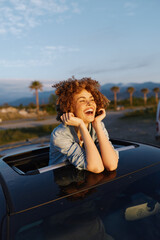 Fototapeta premium A joyful woman leans from a convertible, smiling with hands near her face, enjoying golden hour light. Outdoor lifestyle shot conveying happiness, vacation, travel, and freedom.