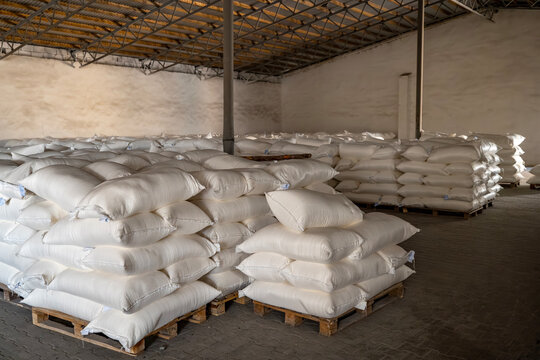 Large quantities of neatly packaged white sacks containing various bulk goods are methodically organized in stacked rows inside a spacious industrial storage facility on wooden pallets