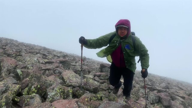 Mountaineer climbs the rocky slopes of Didi Abuli Mountain in Georgia. Hiker uses trekking poles while ascending through fog and harsh weather conditions on a high-altitude volcanic summit.