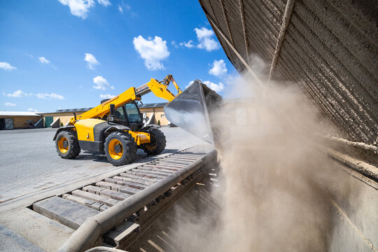 A yellow industrial telehandler operates an articulated boom and bucket, offloading bulk dry aggregate material onto an industrial conveyor system in an outdoor processing facility under a clear sky