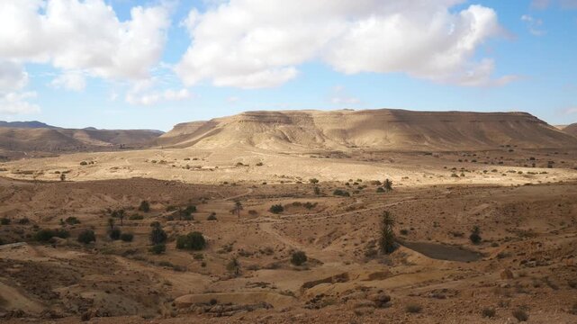 PAN: Expansive desert landscape near Matmata, Tunisia, with rolling arid hills, scattered shrubs, and dramatic cloud shadows creating contrast across the rugged terrain beneath a vast sky