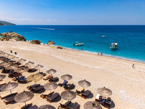 Aerial view of a sandy beach in Sarande with rows of straw umbrellas, sun loungers, turquoise sea, and small boats, Sarande, Vlore County, Albania.