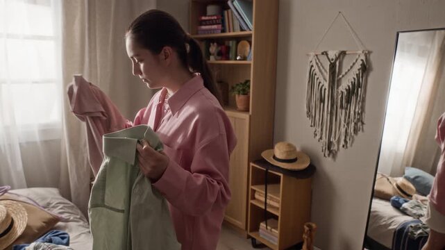 Handheld shot of young Caucasian woman choosing outfits and holding clothes in front of mirror, preparing for travel. Clip is perfect for fashion retail ads, travel planning, and lifestyle vlogs