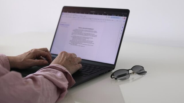 Senior woman working on a laptop document at home with eyeglasses on the table. Close-up scene of online paperwork, digital skills and everyday computer use.