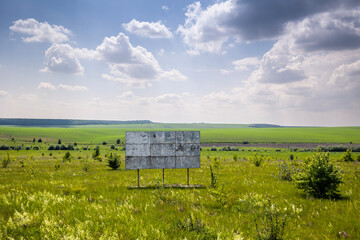 Obraz premium A weathered, blank billboard stands in a vast grassy field with scattered bushes under a partly cloudy sky. Expansive green agricultural fields stretch towards a distant treeline at the horizon