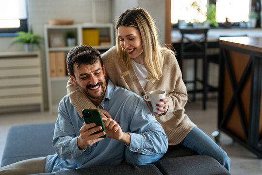 Happy couple enjoying morning coffee at home while browsing their smartphone, showing love