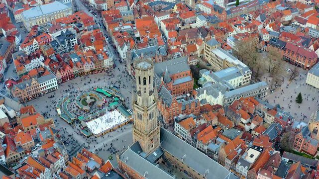 Aerial drone view of Bruges Markt square, Belgium, showing the medieval Belfry tower, Christmas market stalls, and outdoor ice skating rink during winter.