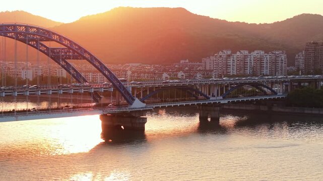 Aerial view of a blue bridge with cars crossing over a river at sunset with a city skyline in the background. Hangzhou, China.
