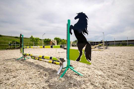 An outdoor equestrian arena with show jumping obstacles and a horse silhouette cutout on a jump wing. The sandy textile riding surface under a bright, cloudy sky in an open field setting