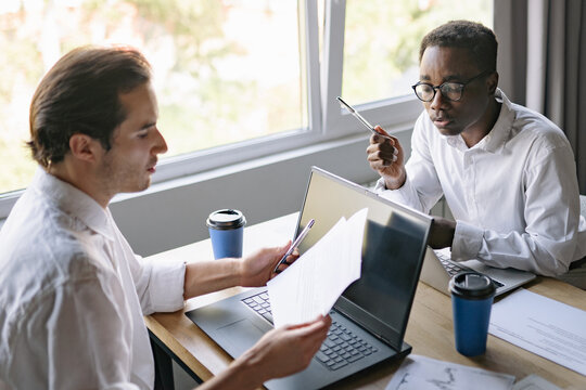 Two mixed-race colleagues are engaged in a discussion over financial documents and laptops at a workspace, focusing on project details and data analysis during the afternoon hours
