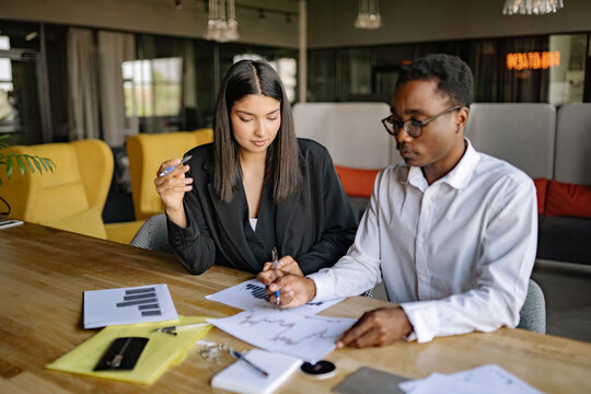 Two professionals, black man in white shirt and Indian woman in suit engage in a focused discussion, analyzing graphs and financial reports on a wooden table in a contemporary office environment.