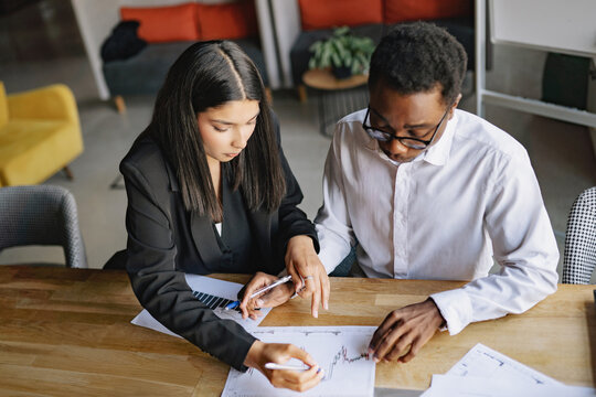 Diverse colleagues analyzing business charts together at a wooden table in a professional office setting during a bright daytime work session