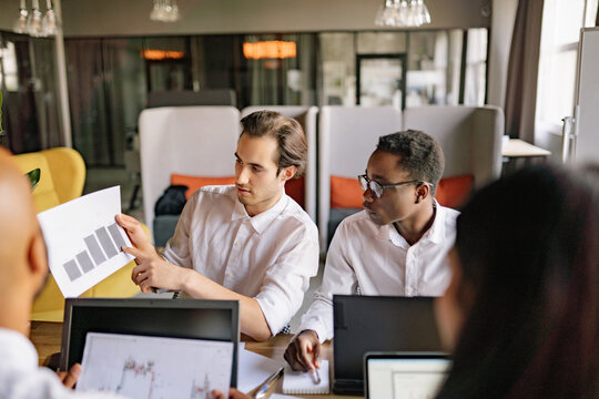 A diverse group of professionals collaborates on a business presentation in a modern office setting, white man shows to the black man financial data insights and strategies during a morning meeting