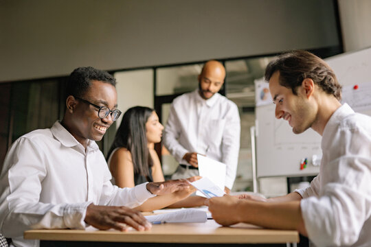 Four diverse colleagues collaborate during a productive daytime meeting in a modern office. Two men sit at a desk reviewing documents, smiling