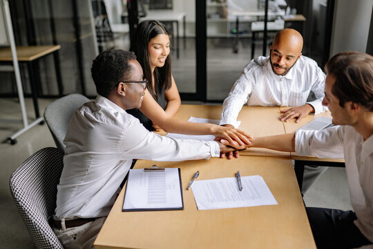 Multiracial colleagues in an office stacking their hands together on a meeting table, demonstrating strong teamwork, unity, and a friendly collaborative spirit