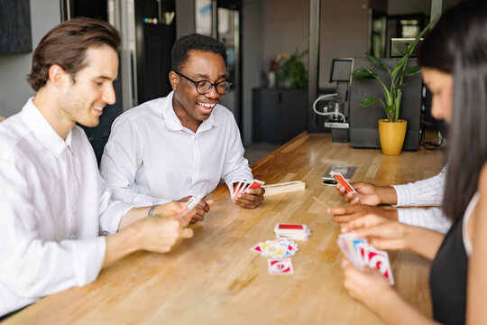 A group of four friends joyfully engages in a card game at a stylish cafe, laughter filling the air as they focus on their hands while sipping beverages and enjoying each other's company