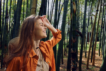 Fototapeta premium Woman portrait in a bamboo forest with a bright smile, wearing a straw hat and orange jacket, sunlight filtering through stalks, nature scene conveying joy and peaceful outdoor exploration.