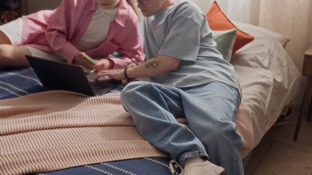 Tilt shot of young adult Caucasian couple sitting on bed, using laptop and smartphone to book vacation next to packed suitcase. Suitable for travel agency promos or online booking platform commercials