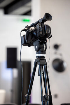 A professional video camera stands on a black tripod, ready for recording an event in an indoor setting. The black camcorder features an eyepiece and various controls, positioned in a room with blurre