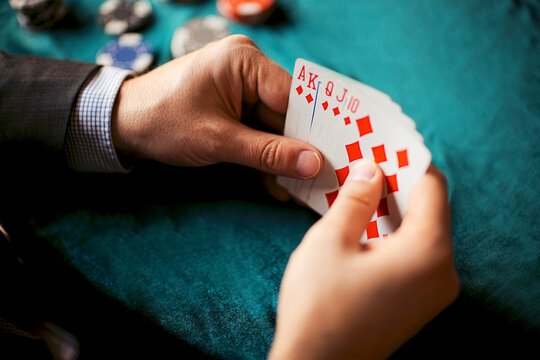 Male gambler holding playing cards with red diamonds and letters AK, surrounded by poker chips on a green felt table in a casino setting