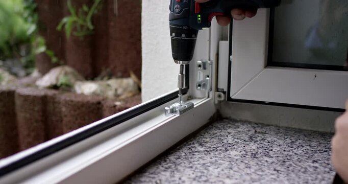 A person uses a cordless electric screwdriver to screw a metal strike plate into a white window frame