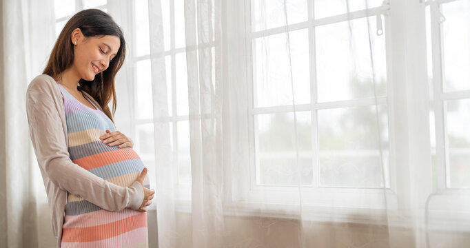 Pregnancy healthcare, beautiful asian pregnant woman holding her belly standing by bright window at home, Happy expecting mother smiling with great love, Maternity lifestyle, sweet newborn preparation