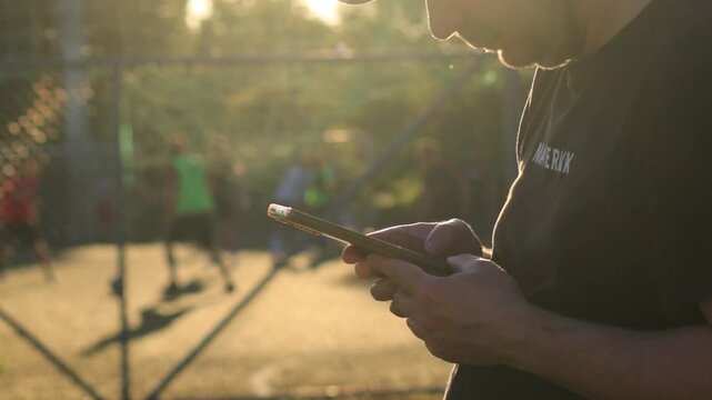 White man using smartphone near football field at sunset while players train in background. Concept of phone addiction, social isolation, procrastination, digital distraction, unhealthy lifestyle