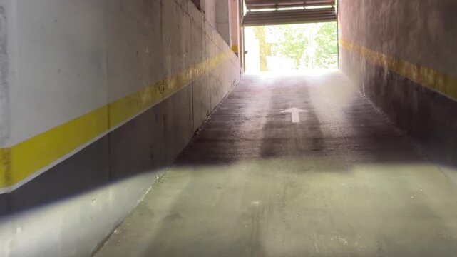 MADRID, SPAIN, June 24-2026: View from inside a parking garage toward daylight. Concrete walls with yellow safety stripes. Road leads to green trees and residential buildings. One parked car visible n