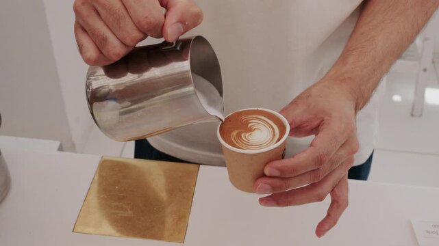 4K Video of a professional barista pouring steamed milk to create beautiful latte art in a paper cup.