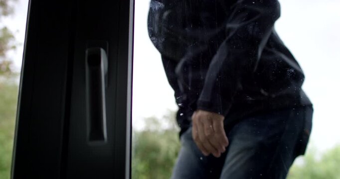 A masked person wearing a black jacket climbs through an open window from the outside