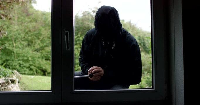 A person in a black hooded jacket and a mask sits outside a window holding a large tool