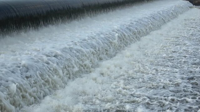 A dynamic, high-velocity shot of chaotic white foam and turbulent currents churning violently in a river downstream from a major water infrastructure discharge.
