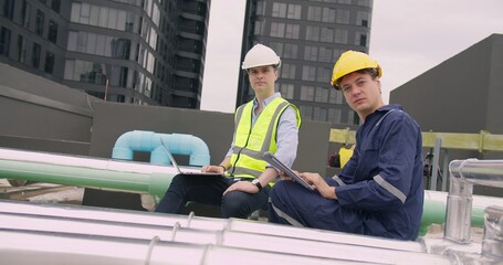 Fototapeta premium Two engineers collaborating on a building rooftop, inspecting the HVAC system and utility infrastructure using a laptop and tablet for site management.