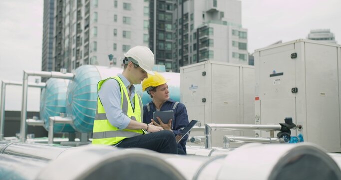 Two industrial engineers collaborate on system diagnostics, using a laptop and tablet to inspect a large HVAC installation on a building rooftop.