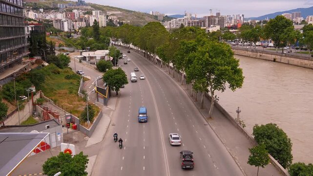 Aerial view of cars and vehicles driving along a multi-lane highway next to the mtkvari river, with the modern cityscape of tbilisi, georgia, expanding into the hillside in the background