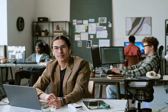 Portrait of young adult Asian man working at laptop in modern office, leading digital team and looking at camera. Ideal for startup marketing, teamwork, productivity, tech workplace