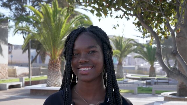 Portrait of a confident young black woman with braids smiling at the camera in a sunny city park