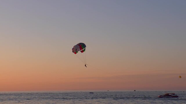 Single person parasailing over the calm sea during a beautiful orange sunset, representing an adventurous summer vacation activity with a speedboat pulling the colorful parachute through the air