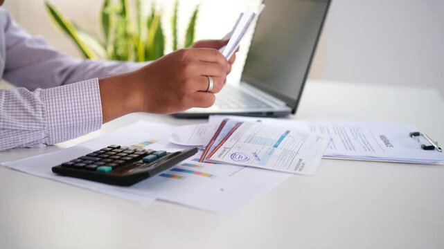 An accountant holds receipts or invoices, checks business documents, passbooks, bank statements, and calculates expenses paid via credit card before completing the bookkeeping.
