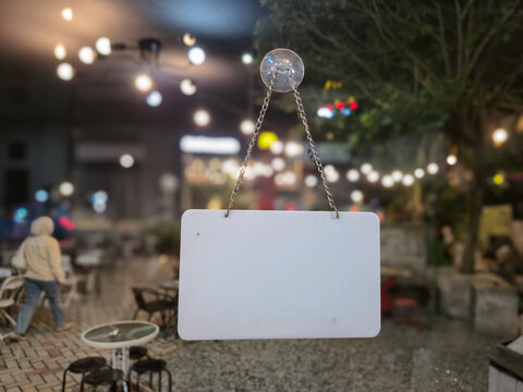 ​Blank white hanging sign mockup with chain on glass window, blurred cozy cafe restaurant background with warm bokeh lights, copy space for text or logo