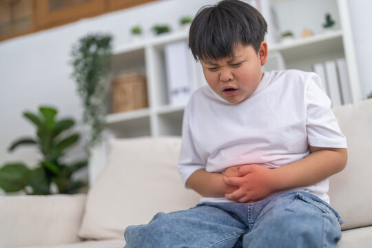 Portrait of sick unhappy little child asian boy touching belly stomach with suffering from stomach ache pain, gastritis, gastroenteritis, food poisoning, diarrhea, intestinal inflammation at home