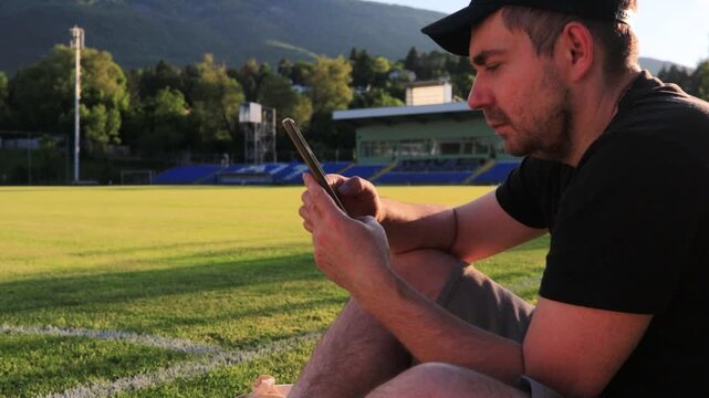 A white man sits relaxing and unwinding on the green grass of a football stadium at sunset, browsing his mobile phone. Concept of leisure time, summer evening, sports field, urban recreation, outdoor