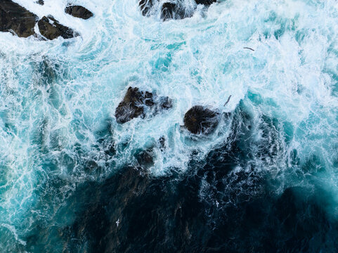Aerial view of ocean waves crashing over dark coastal rocks