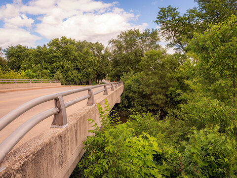 Concrete road bridge with metal guardrails crossing over a river surrounded by lush green trees and foliage under a cloudy blue sky