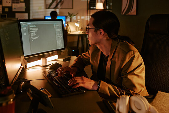 Asian man coding at desktop computer in office, working late on software development and cybersecurity tasks. Side view, fitting tech team, IT service, remote work marketing