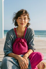 Fototapeta premium Seated woman portrait with a magenta knit bag and relaxed grey outfit on a sunlit pier for lifestyle banner and fashion editorial use