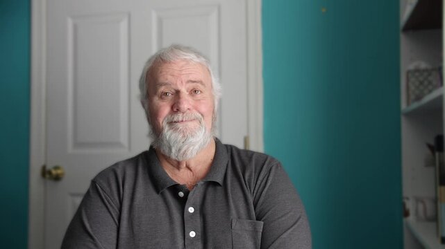 An elderly man sits in a room with a blue wall. He smiles and waves. The lighting is bright. He interacts with the camera and expresses warmth. This moment shows connection and friendliness.